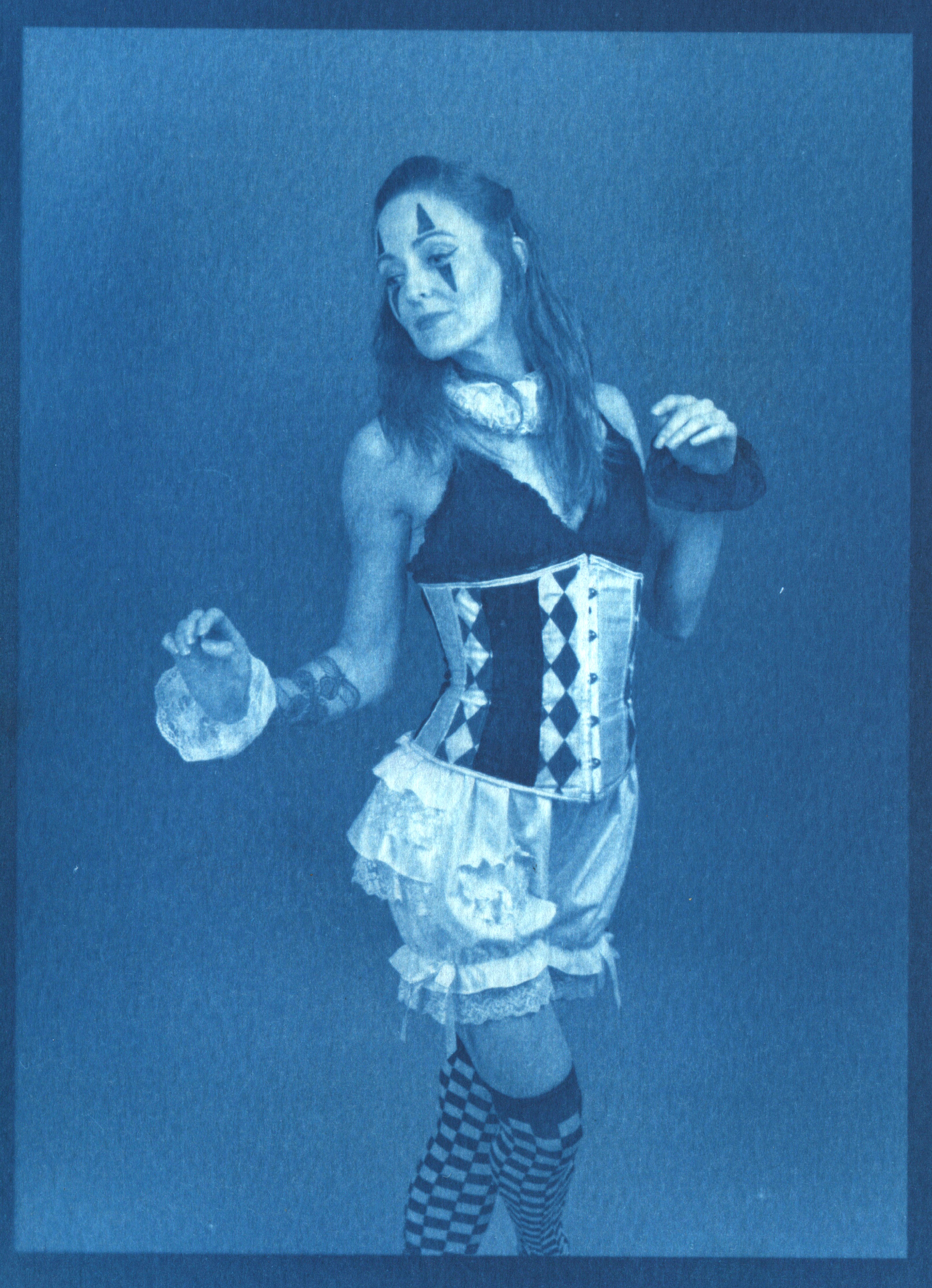 Cyanotype print of Laura in a harlequin circus costume — a diamond-patterned corset over a ruffled skirt with checkerboard stockings — posed with hands raised and painted clown-like face markings