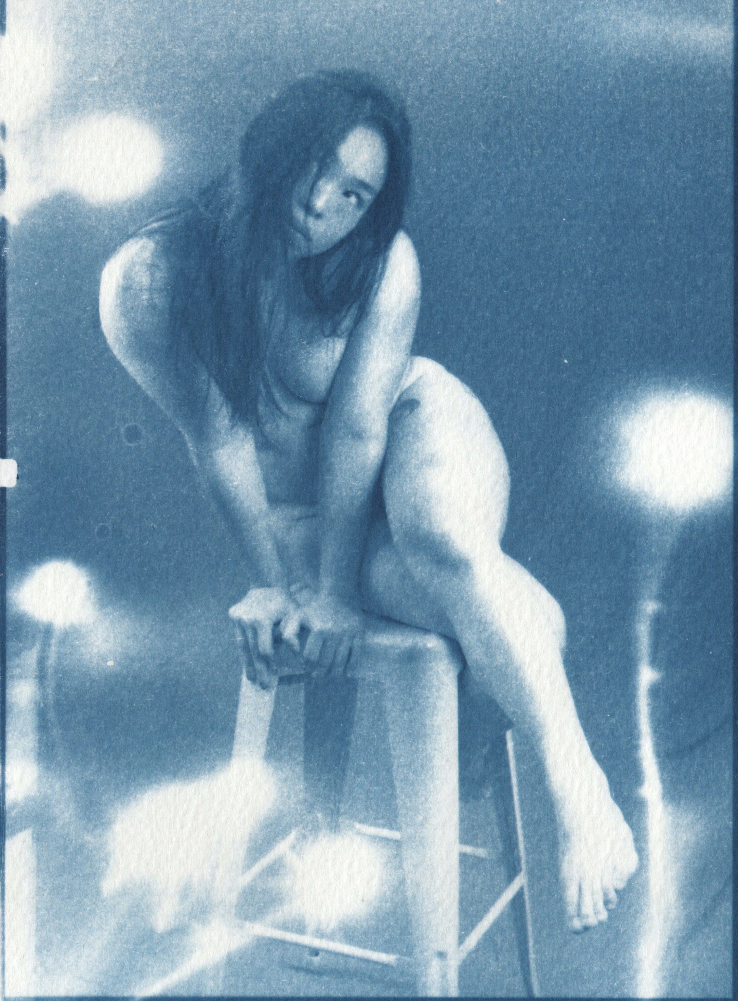 Cyanotype print of Lorraine seated on a stool, leaning forward with legs crossed, surrounded by botanical photograms of plants